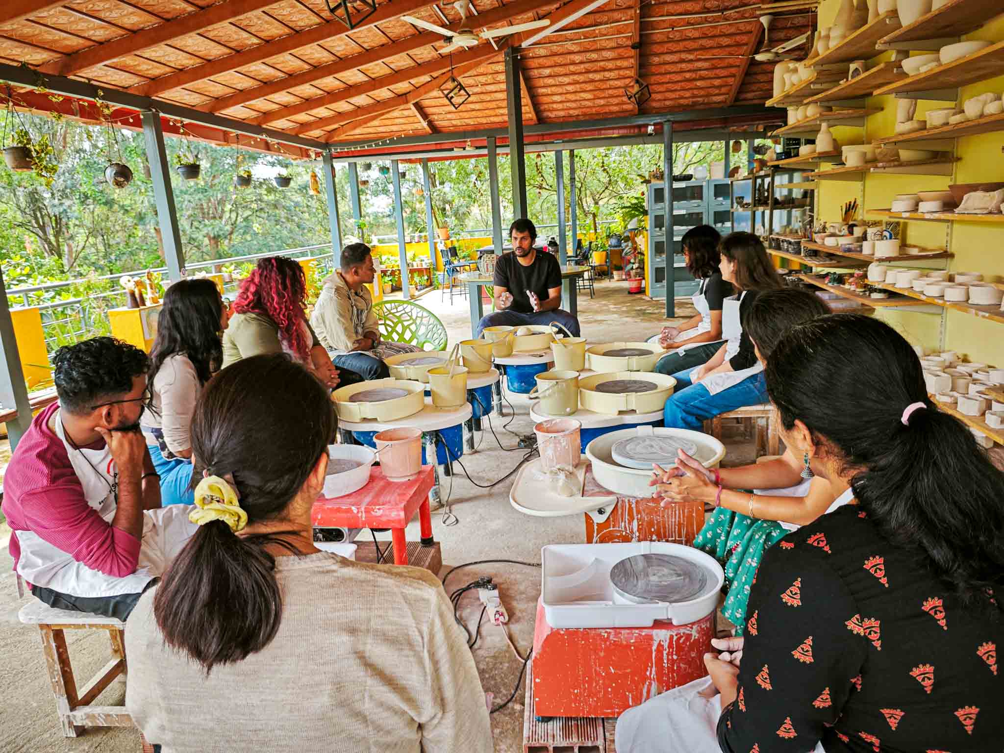 Group of pottery learners sitting around pottery wheels receiving instruction in an open-air studio.
